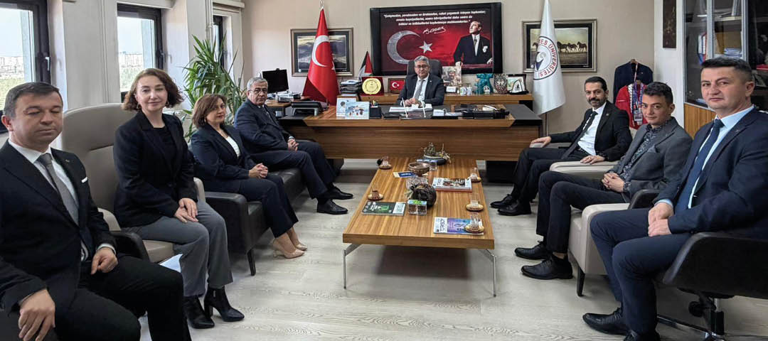 A group of people in formal attire are seated in an office around a large desk with flags, plaques, and a framed portrait of a historical figure with a flag in the background. Description generated by AI
