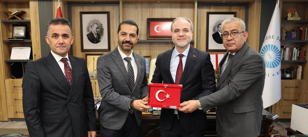Four people in suits stand indoors holding a red plaque with a white crescent and star symbol, with framed pictures and a flag in the background. Description generated by AI