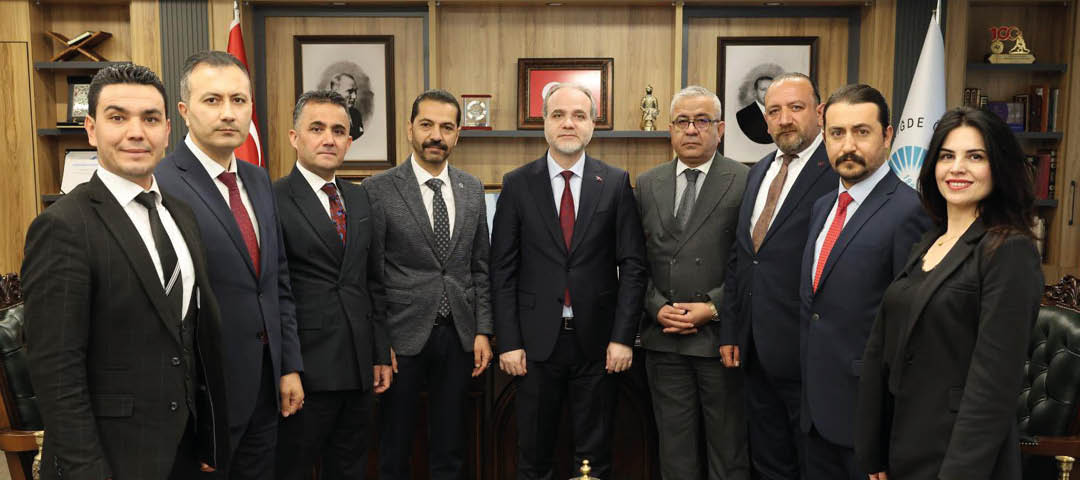 Seven adults dressed in formal suits stand closely together in an office setting with framed portraits and decorative items on wooden shelves behind them. Description generated by AI