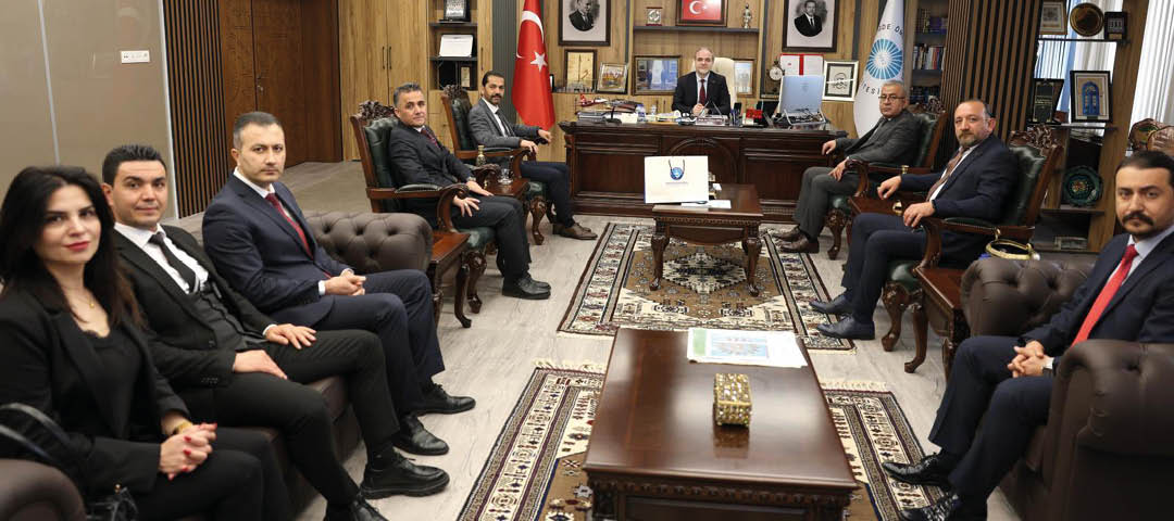 Several people in formal attire are seated in a wood-paneled office with flags, portraits, and a large desk where one person is sitting behind it. Description generated by AI